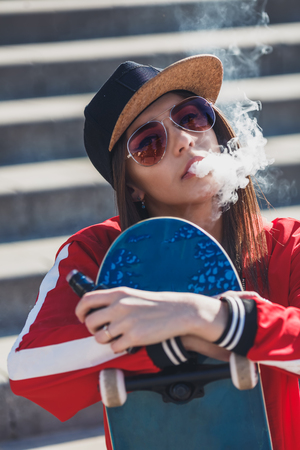 Vaping girl. Young woman with skateboard vape e-cig. Pretty young female in black hat, red clothing vape ecig, vaping device at the sunset. Toned image. Hip-hop style.の写真素材