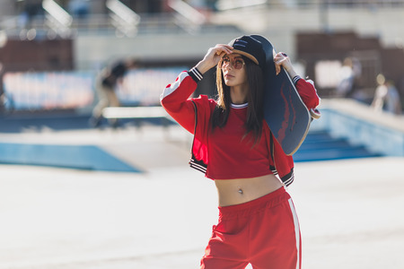 Portrait of young female wearing black hat, red clothing skateboarder holding her skateboard. Woman with skating board looking at camera outdoors.の写真素材