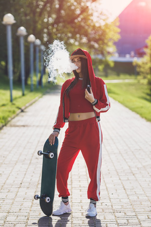 Vaping girl. Young woman with skateboard vape e-cig. Pretty young female in black hat, red clothing vape ecig, vaping device at the sunset. Toned image. Hip-hop style.の写真素材