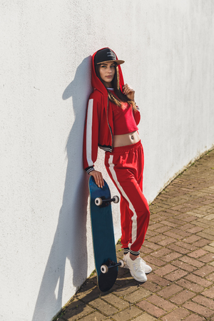 Portrait of young female wearing black hat, red clothing skateboarder holding her skateboard. Woman with skating board looking at camera outdoors.の写真素材