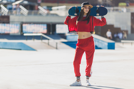 Portrait of young female wearing black hat, red clothing skateboarder holding her skateboard. Woman with skating board looking at camera outdoors.の写真素材