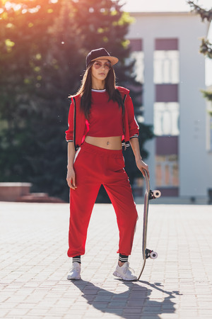 Portrait of young female wearing black hat, red clothing skateboarder holding her skateboard. Woman with skating board looking at camera outdoors.の写真素材