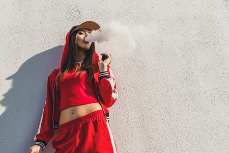 Vaping girl. Young woman standing in front of a white wall and vape e-cig. Pretty young female in black hat, red clothing vape ecig, vaping device at the sunset. Toned image. Hip-hop style.の写真素材