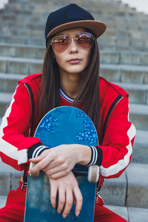 Portrait of young female wearing black hat, red clothing skateboarder holding her skateboard. Woman with skating board looking at camera outdoors.の写真素材