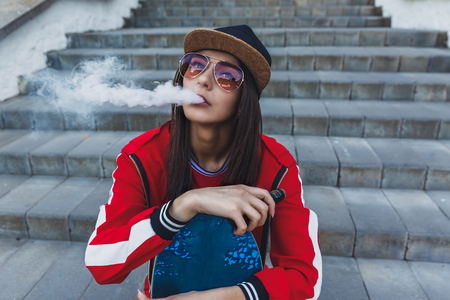 Vaping girl. Young woman with skateboard vape e-cig. Pretty young female in black hat, red clothing vape ecig, vaping device at the sunset. Toned image. Hip-hop style.の写真素材