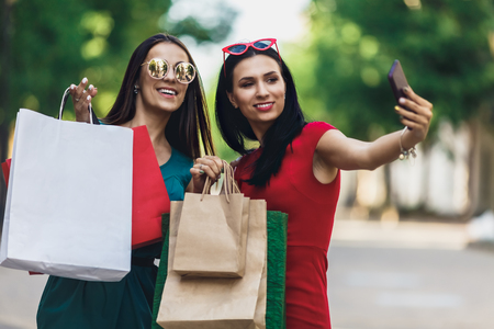 Beautiful happy girls in sun glasses holding shopping bags, making selfie on smart phone and smiling. Sale and consumerism concept.の写真素材