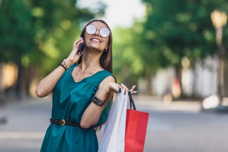 Close up of young charming dark-haired caucasian female with shopping bags smiling with teeth and talking on phone. Positive emotions and Shopping day concept.の写真素材