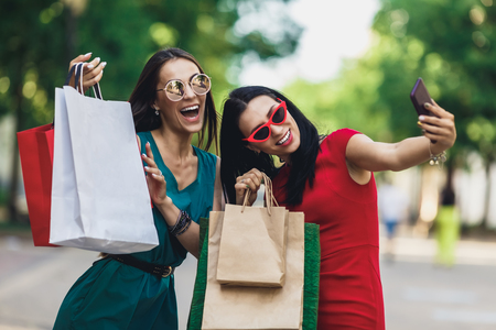 Beautiful happy girls in sun glasses holding shopping bags, making selfie on smart phone and smiling. Sale and consumerism concept.の写真素材