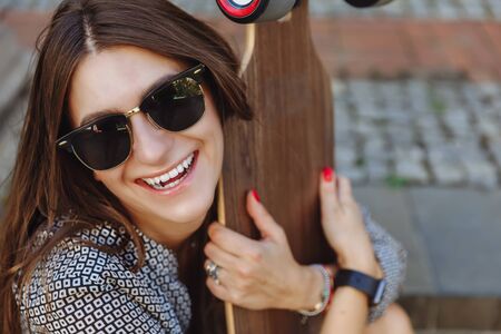 Close up Portrait of Beautiful young woman sitting on the ground and holding a longboard in the city street sunny weather. Young hipster girl posing with longboard, skateboard, streetphoto, life style, freedom, happy face.の写真素材