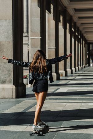 Beautiful young skater woman riding on her longboard in the city. Stylish girl in street clothes...