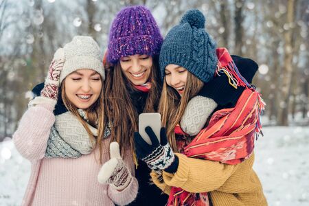 Three women friends outdoors in knitted hats using mobile phone on a snowy cold weather. Group of young female friends outdoors in winter park.の写真素材
