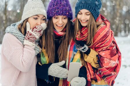 Three women friends outdoors in knitted hats using mobile phone on a snowy cold weather. Group of young female friends outdoors in winter park.の写真素材