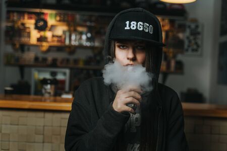 Young pretty woman in cap smoke an electronic cigarette at the vape shop. Hip-hop style. Closeup.の写真素材