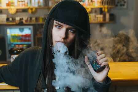 Young pretty woman in cap smoke an electronic cigarette at the vape shop. Hip-hop style. Closeup.の写真素材