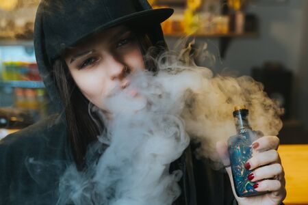 Young pretty woman in cap smoke an electronic cigarette at the vape shop. Hip-hop style. Closeup.の写真素材