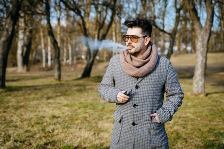 Confident young bearded male in sunglasses vape electronic cigarette. A young handsome bearded hipster man vaping e-cigarette outdoors in the park. Close up.の写真素材