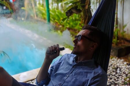 Man resting in a hammock and vaping and letting off steam from an electronic cigaretteの写真素材