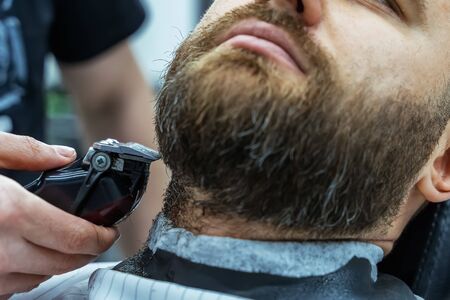 Barber trimming bearded man with shaving machine in barbershop. Hairstyling process. Close-up of a Hairstylist cutting the beard of a bearded male.の写真素材