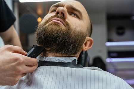 Barber trimming bearded man with shaving machine in barbershop. Hairstyling process. Close-up of a Hairstylist cutting the beard of a bearded male.の写真素材