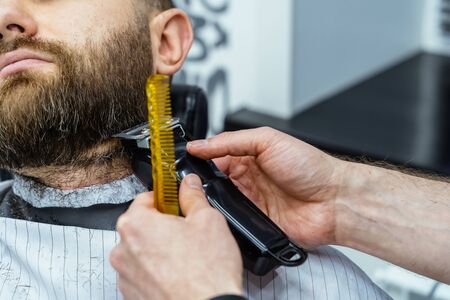 Barber trimming bearded man with shaving machine in barbershop. Hairstyling process. Close-up of a Hairstylist cutting the beard of a bearded male.の写真素材