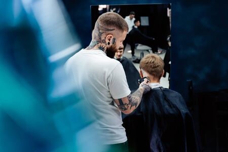 Male haircut with electric razor. Tattooed Barber makes haircut for client at the barber shop by...