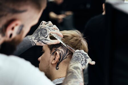 Tattooed Barber cuts the hair of the client with scissors. Close up. Attractive male is getting a modern haircut in barber shop.の写真素材