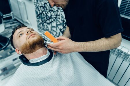 Barber trimming bearded man with shaving machine in barbershop. Hairstyling process. Close-up of a Hairstylist cutting the beard of a bearded male.の写真素材