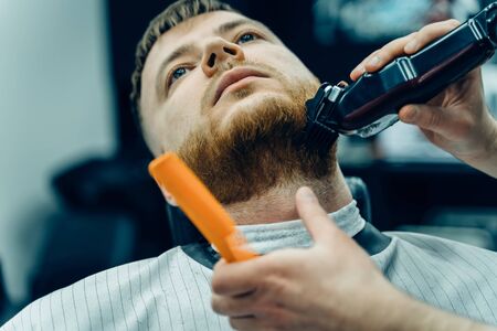 Barber trimming bearded man with shaving machine in barbershop. Hairstyling process. Close-up of a Hairstylist cutting the beard of a bearded male.の写真素材