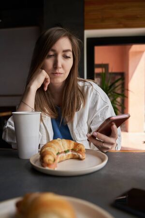 Front view of attractive woman wearing glasses using smartphone, drinking coffee in cafe. Female texting and sharing messages on social media, enjoying mobile technology, relaxing in coffee shop.の写真素材