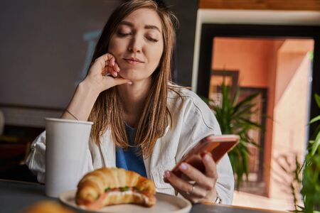 Front view of attractive woman wearing glasses using smartphone, drinking coffee in cafe. Female...