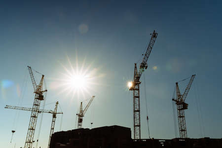 Industrial construction cranes and building silhouettes over sun at sunrise or sunset.