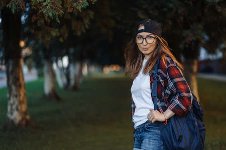 Happy Young female student dressed in casual clothing with cup of coffee and backpack behind her back walking around city. Woman student holding laptop and drinking coffee. Summer sunset time.の写真素材