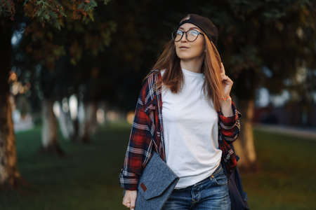 Happy Young female student dressed in casual clothing with cup of coffee and backpack behind her back walking around city. Woman student holding laptop and drinking coffee. Summer sunset time.の写真素材