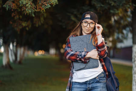 Happy Young female student dressed in casual clothing with cup of coffee and backpack behind her back walking around city. Woman student holding laptop and drinking coffee. Summer sunset time.の写真素材