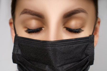 Eyelash Extension Procedure. Beautiful Woman with Extreme Long False Eyelashes. Makeup, Cosmetics. Beauty, Skincare. Woman in medical mask.の写真素材
