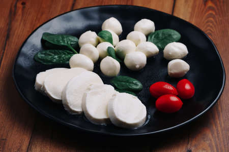 White small mozzarella cheese balls, spinach leaves and tomatoes on black plate.の写真素材
