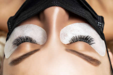 Eyelash Extension Procedure. Beautiful Woman with Extreme Long False Eyelashes. Makeup, Cosmetics. Beauty, Skincare. Woman in medical mask.の写真素材