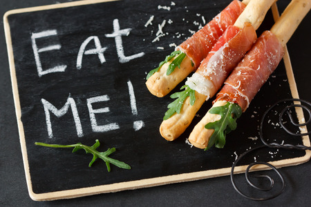 Grissini bread sticks with prosciutto ham, arugula and parmesan cheese on a black chalkboard.の写真素材