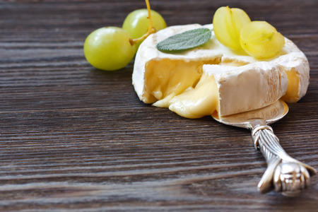 Round Camembert cheese with a section cut out and served grape and leaf of sage.の写真素材