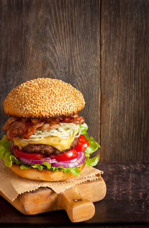 Big delicious cheeseburger stacked high with a bacon, beef patty, coleslaw, cheese, lettuce, red onion and tomato on sesame seed bun served  on wooden cutting board.の写真素材
