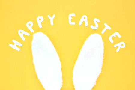 Happy Easter lettering and white bunny fur ears on yellow background. Holiday composition creative concept. Idea for postcard, banner, poster. Top view, flatlayの写真素材