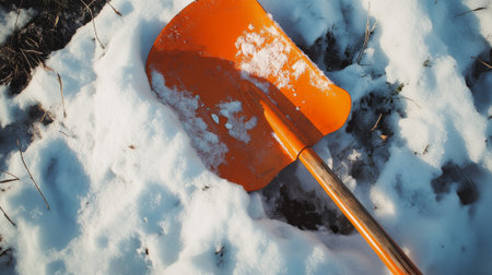 Orange snow shovel in snow. A bright orange snow shovel lies partially buried in a snowdrift, with patches of bare ground and dried grass visible in a sunny outdoor setting.の素材
