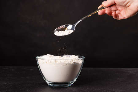 Female hand sprinkles flour from a spoon into a bowl on a black background