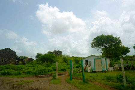 This image portrays an abandoned rural scene, showcasing deteriorating buildings shrouded in overgrown plant life. Prominently in the foreground is a weathered stone structure with a roof that has partially fallen in, accompanied by vegetation that climbs its walls. A neglected rusty cart rests on the grassy area, enhancing the feeling of desolation. In the background, additional rundown buildings with red-tiled roofs and a larger stone structure can be discerned. The clear sky, with a few clouds scattered throughout, serves to amplify the contrast between the idyllic landscape and the decaying structures.の写真素材