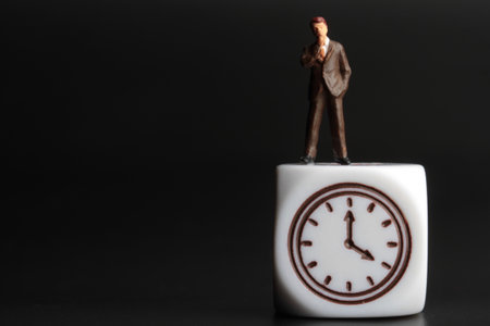 Time management concept. Miniature figurine of a businessman standing on a white cube clock. Black background.の写真素材
