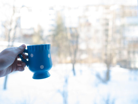 Close up hands of holding a cup of hot cocoa with marshmallow or coffee indoor near window on snow blue backgroundの写真素材