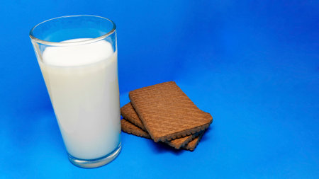 a glass of milk is next to a chocolate cookie on a blue backgroundの写真素材