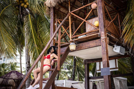 Beautiful and sexy lifeguard in a red bikini lying relaxed on the watchtower looking out for the swimmers, while watching the beautiful turquoise Caribbean Sea and caressing her black hair in the tropical breeze. Baywatch.の写真素材