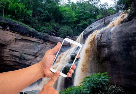 Man hand is holding a modern touch screen phone of waterfall in backgroundの写真素材