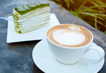Latte or cappuccino coffee in white cup with green tea cake on table